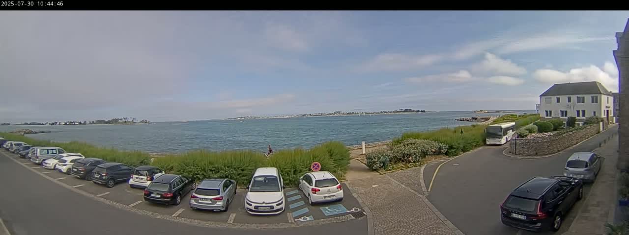 A partly cloudy day reveals a calm body of water with a distant shoreline, several cars parked in a lot near a building and a bus, and a grassy area separating the parking lot from the water.