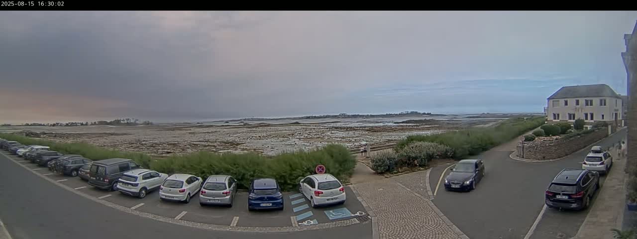 A partly cloudy day reveals a calm body of water with a distant shoreline, several cars parked in a lot near a building and a bus, and a grassy area separating the parking lot from the water.