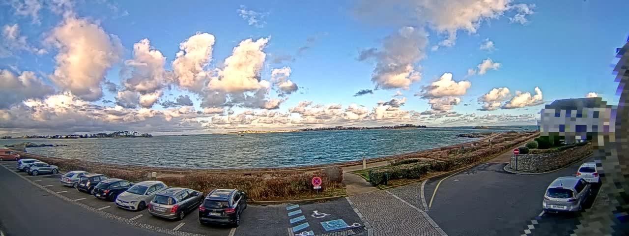 A vibrant, partly cloudy day illuminates a coastal scene where cars are parked along a stone-walled promenade beside a large expanse of choppy blue water, with distant land and buildings visible under a sky full of fluffy white and grey clouds.