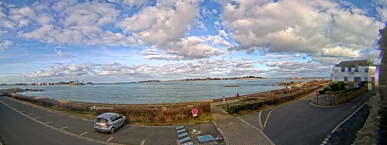 A wide panoramic view captures a bright, partly cloudy sky over a calm bay dotted with distant islands, a winding coastal road, and a silver car parked near lush hedges.