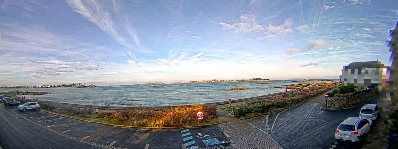 A wide panoramic view captures a bright, partly cloudy sky over a calm bay dotted with distant islands, a winding coastal road, and a silver car parked near lush hedges.