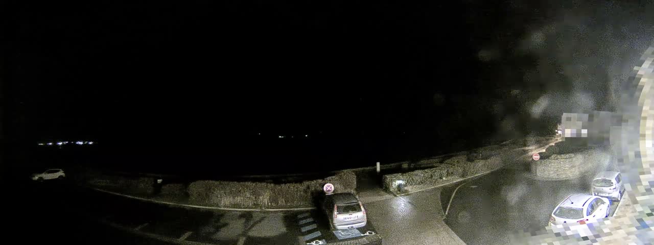 A wide panoramic view captures a bright, partly cloudy sky over a calm bay dotted with distant islands, a winding coastal road, and a silver car parked near lush hedges.