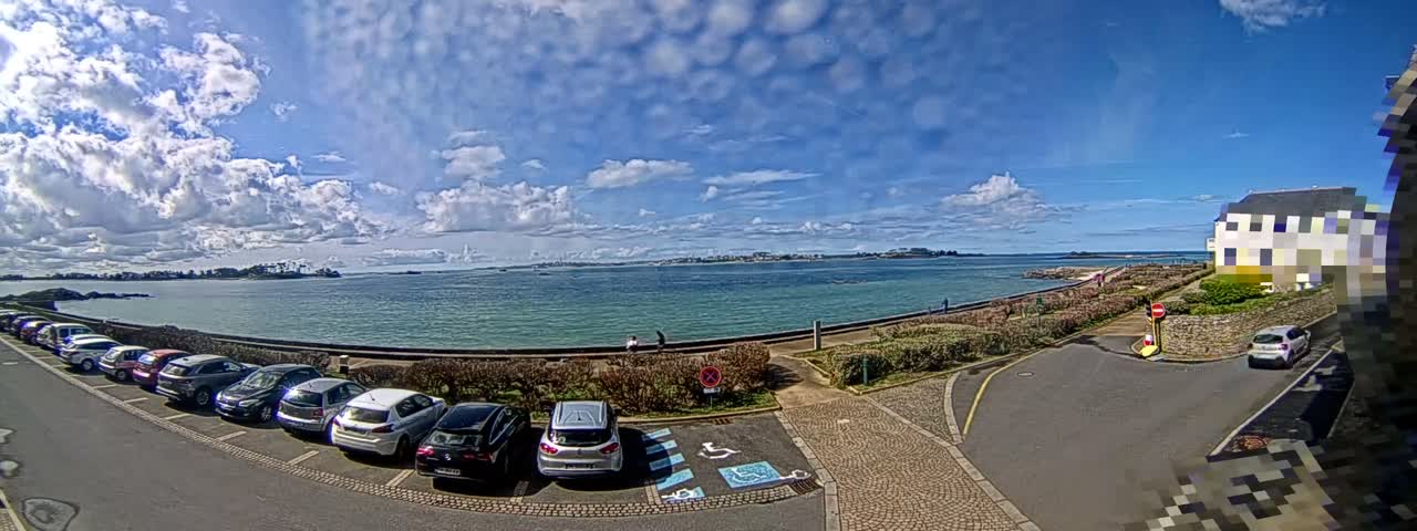 A wide panoramic view captures a bright, partly cloudy sky over a calm bay dotted with distant islands, a winding coastal road, and a silver car parked near lush hedges.