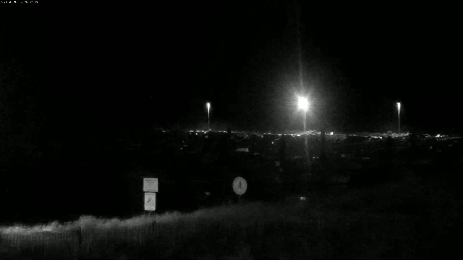 A nighttime, low-light view shows a dark, grassy area in the foreground with a few signs, and in the background are several bright lights illuminating a parking lot or field of vehicles.