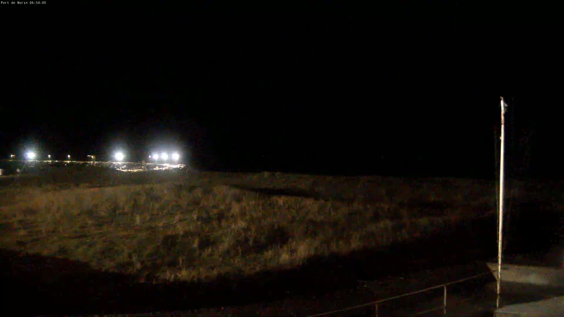 A dark, nighttime scene shows a flat expanse of land with a tall pole on the right, illuminated by distant lights in the background.