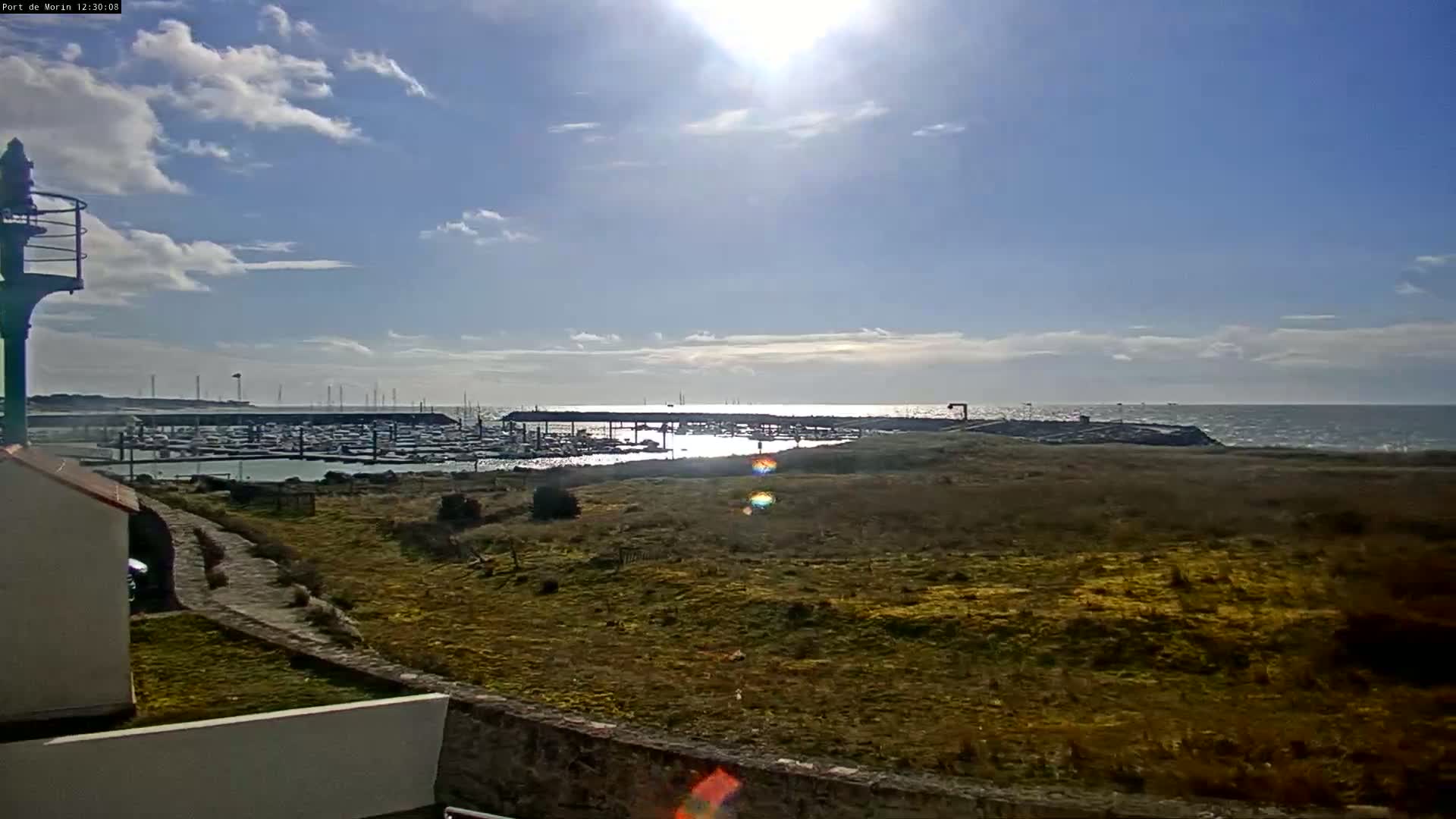 A sunny day with partly cloudy skies shows a marina filled with boats, viewed from a grassy area next to a stone wall and a small lighthouse.
