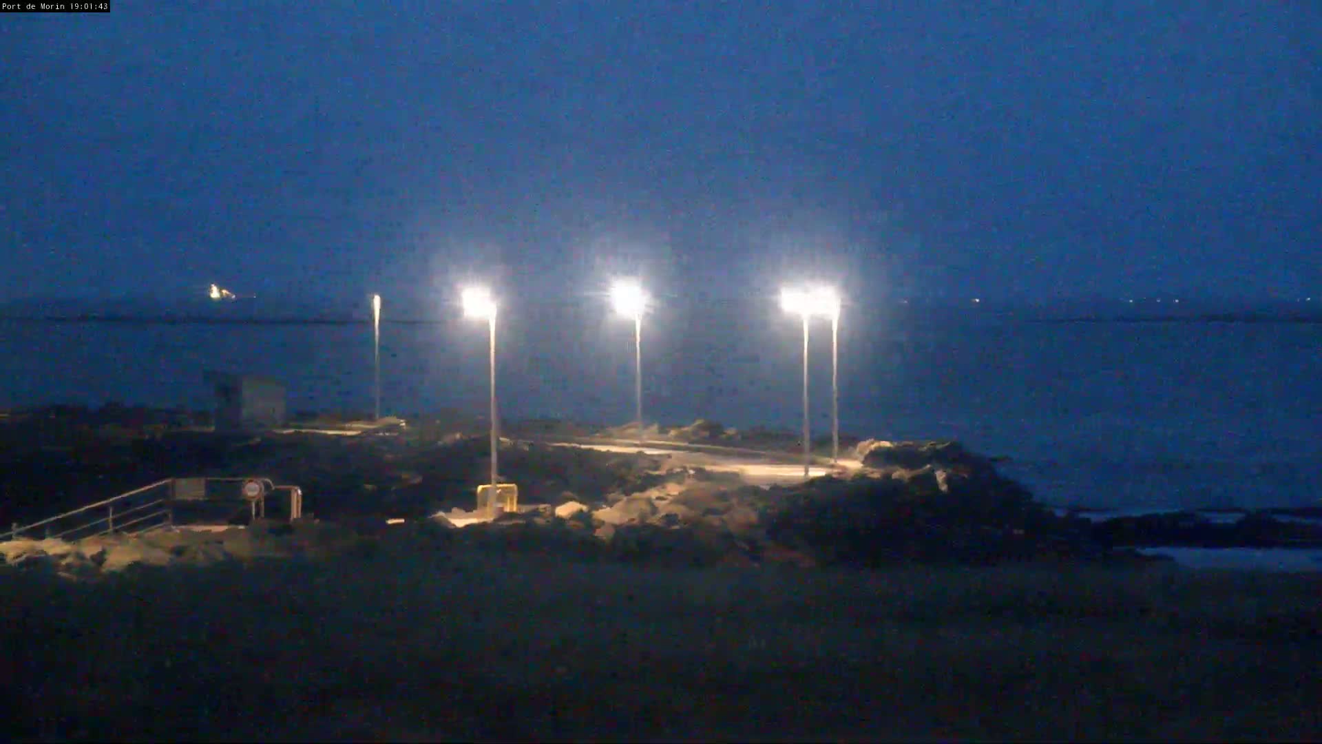 Three illuminated lampposts stand on a rocky shoreline overlooking a dark, calm body of water under a twilight sky.
