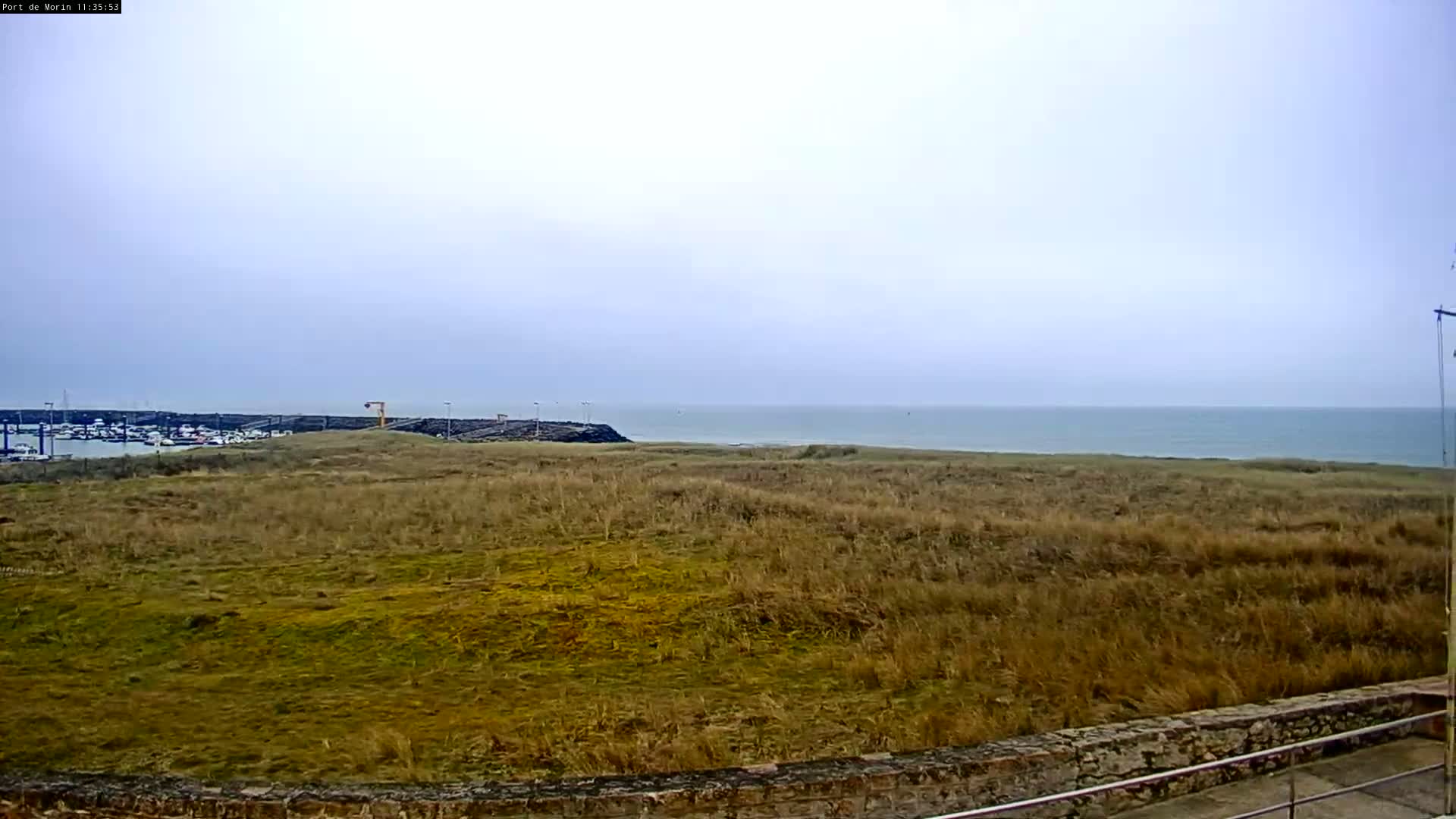 A mostly cloudy day reveals a view of a calm sea beyond a stretch of dry, brown grass leading to a harbor with several boats.