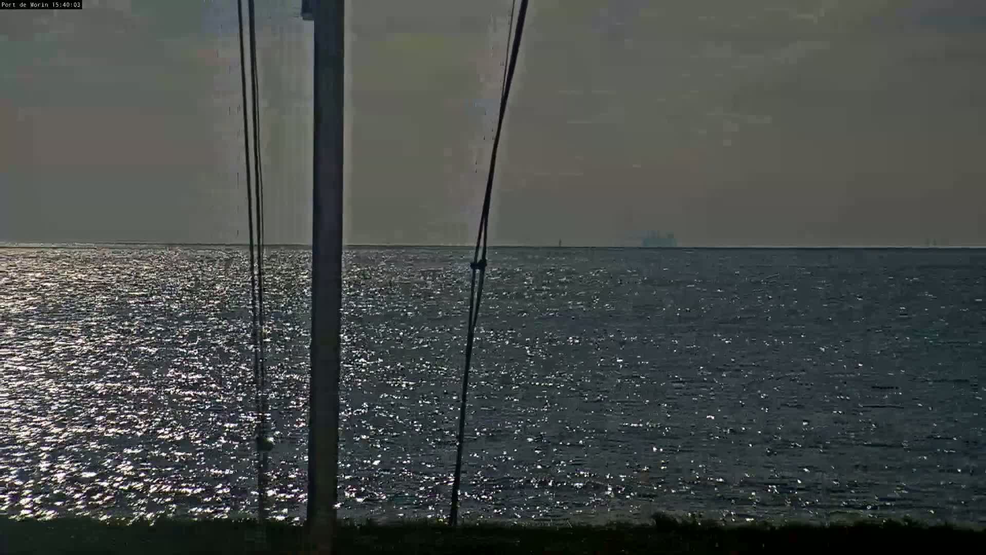 A view of a calm, glistening sea under a mostly cloudy sky, with a distant ship and a vertical pole with ropes in the foreground.