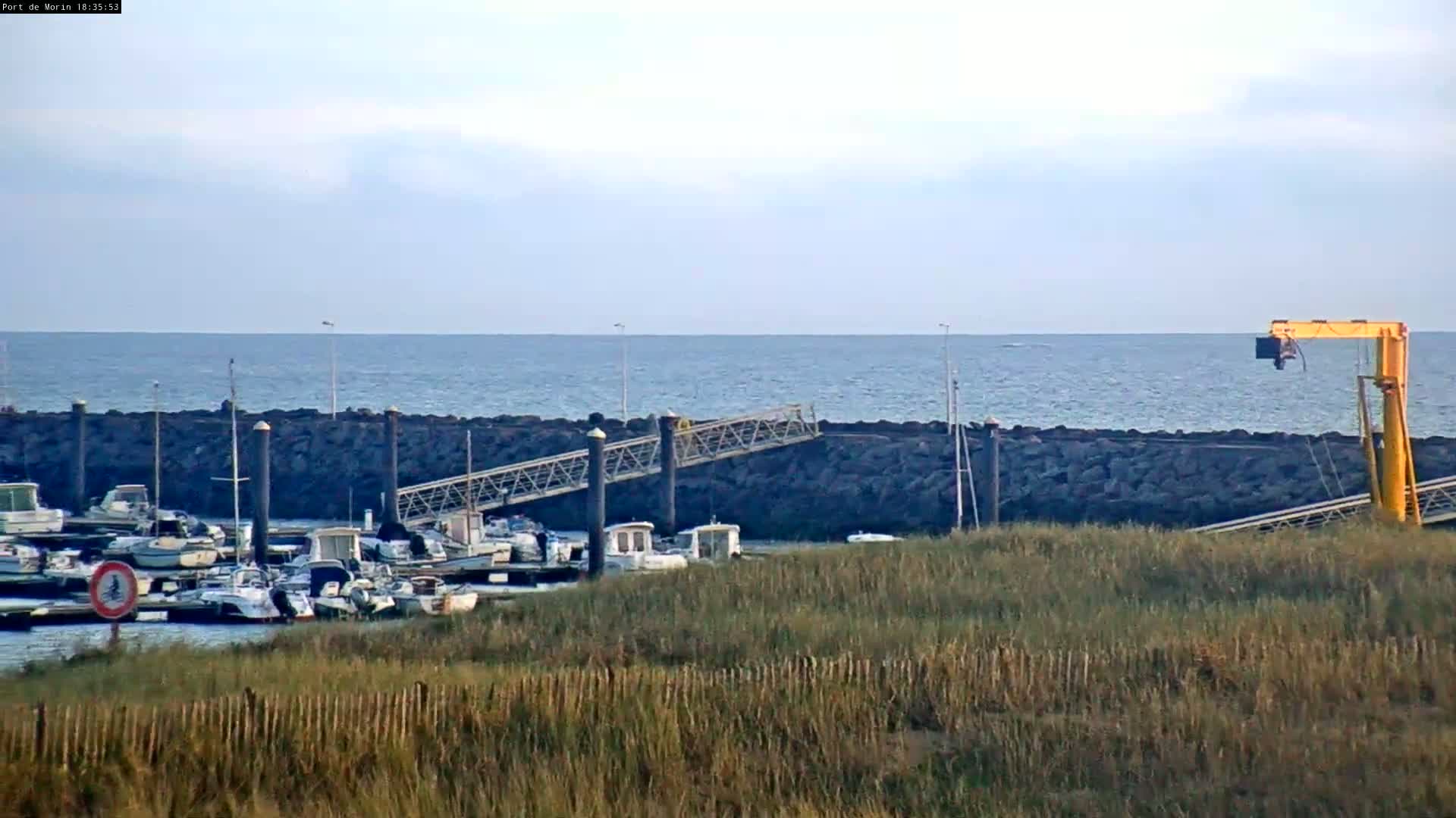 A small harbor with numerous boats is situated along a rocky shore, overlooking a calm sea under a mostly clear sky.