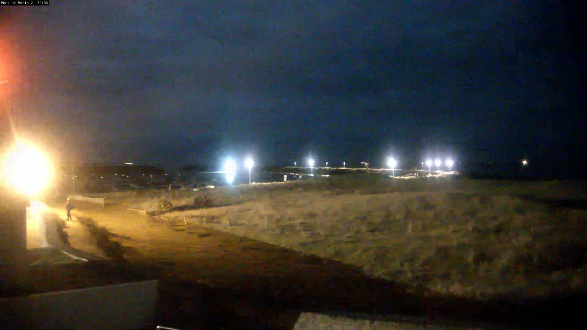 A nighttime view of a sparsely populated coastal area with several lights illuminating a sandy expanse near the water under a dark, clear sky.