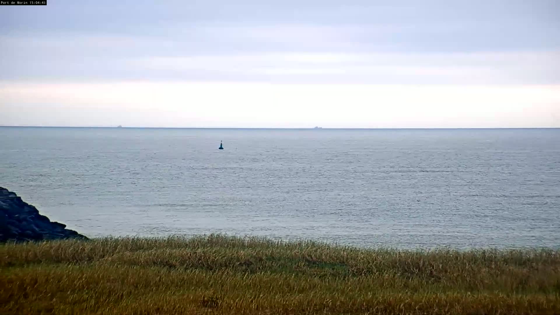 A calm, overcast day reveals a wide expanse of sea with a small buoy and distant ships, viewed from a grassy shoreline with a dark rocky outcrop in the foreground.