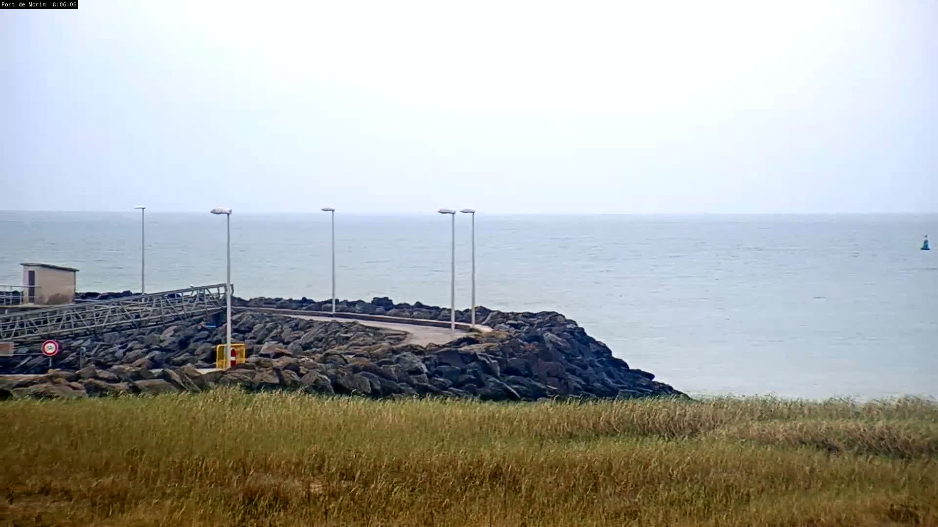 A rocky shoreline with a small walkway and lights overlooks a calm, light-grey sea under an overcast sky.