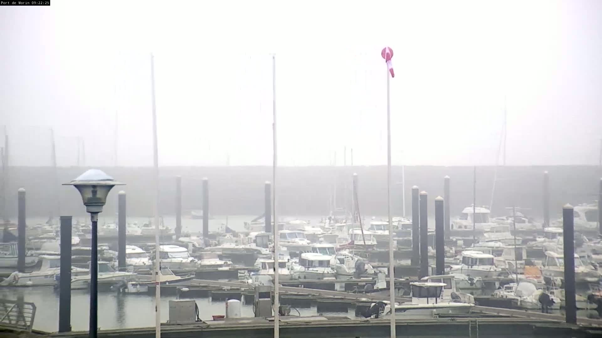 A foggy marina with numerous boats docked at various slips, a windsock, and a lamppost.