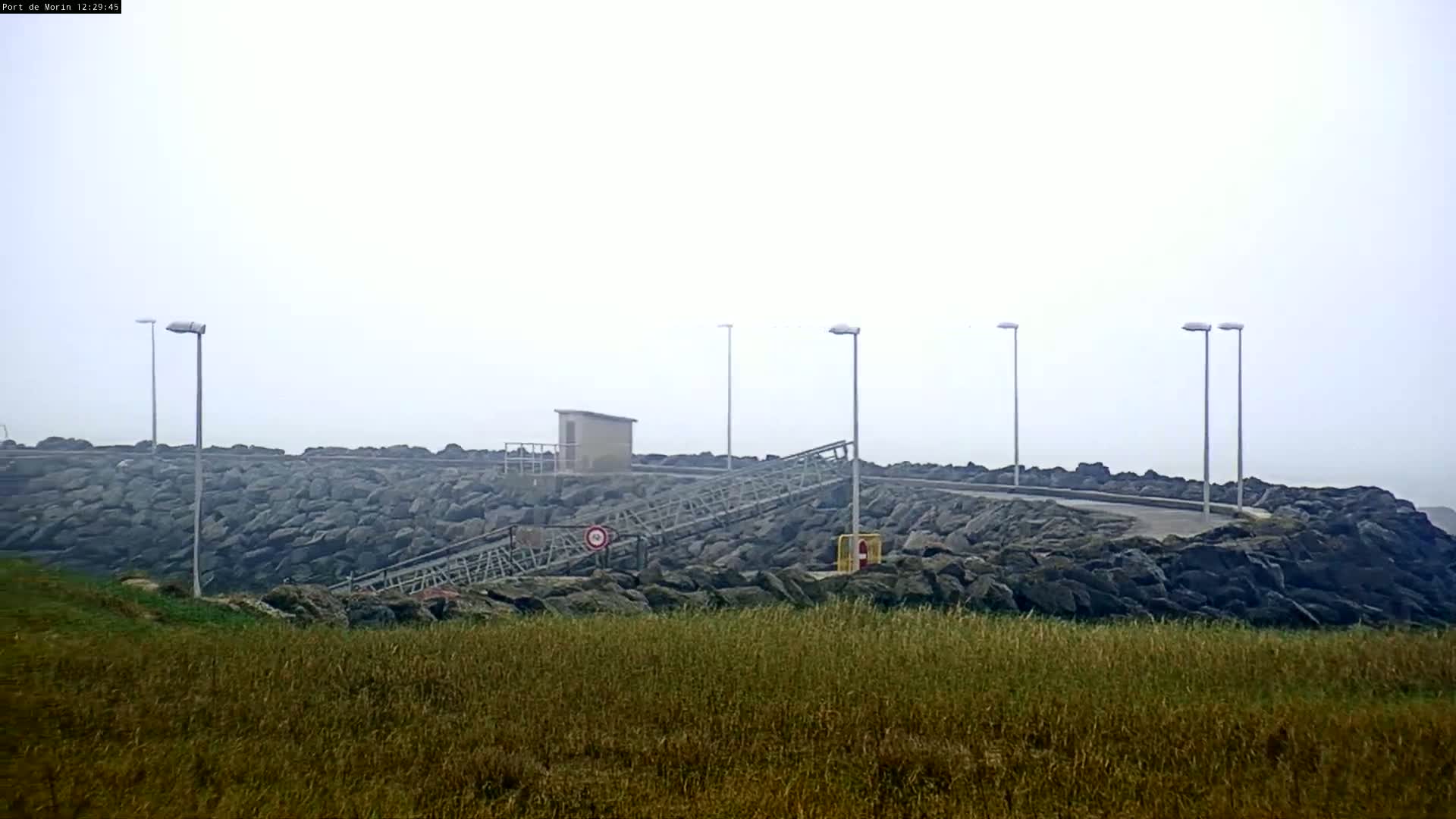 A foggy coastal scene shows a rocky outcrop with several tall lampposts, a small building, and a metal ramp leading down to the water's edge, all situated above a field of tall grass.