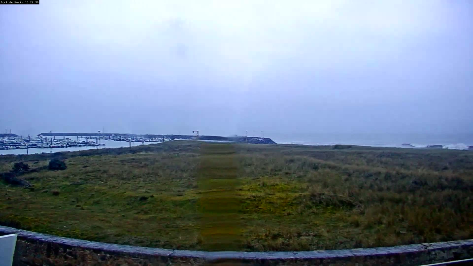 A grassy field with a path leading to a harbor full of boats, under an overcast sky with a rough sea in the background.