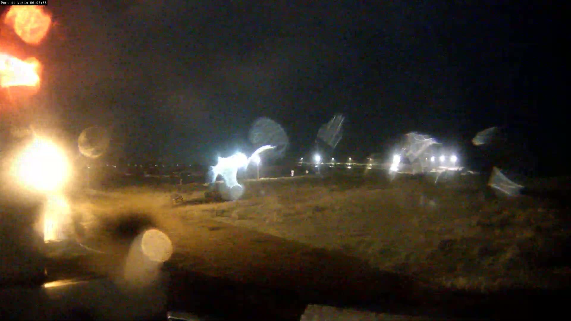 A nighttime, rain-streaked view shows a dimly lit, flat, empty expanse of land with a few distant lights and blurred vehicles.