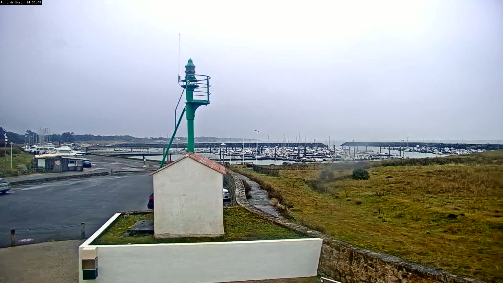 A green light tower stands near a small building overlooking a marina filled with boats under an overcast sky.
