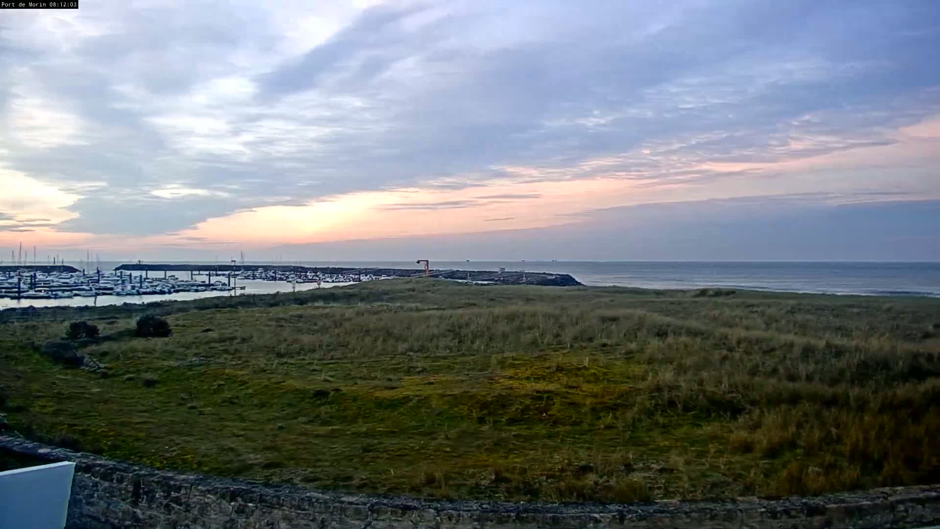 A calm ocean with a marina filled with boats is visible at sunrise under a partly cloudy sky, viewed from a grassy hill.