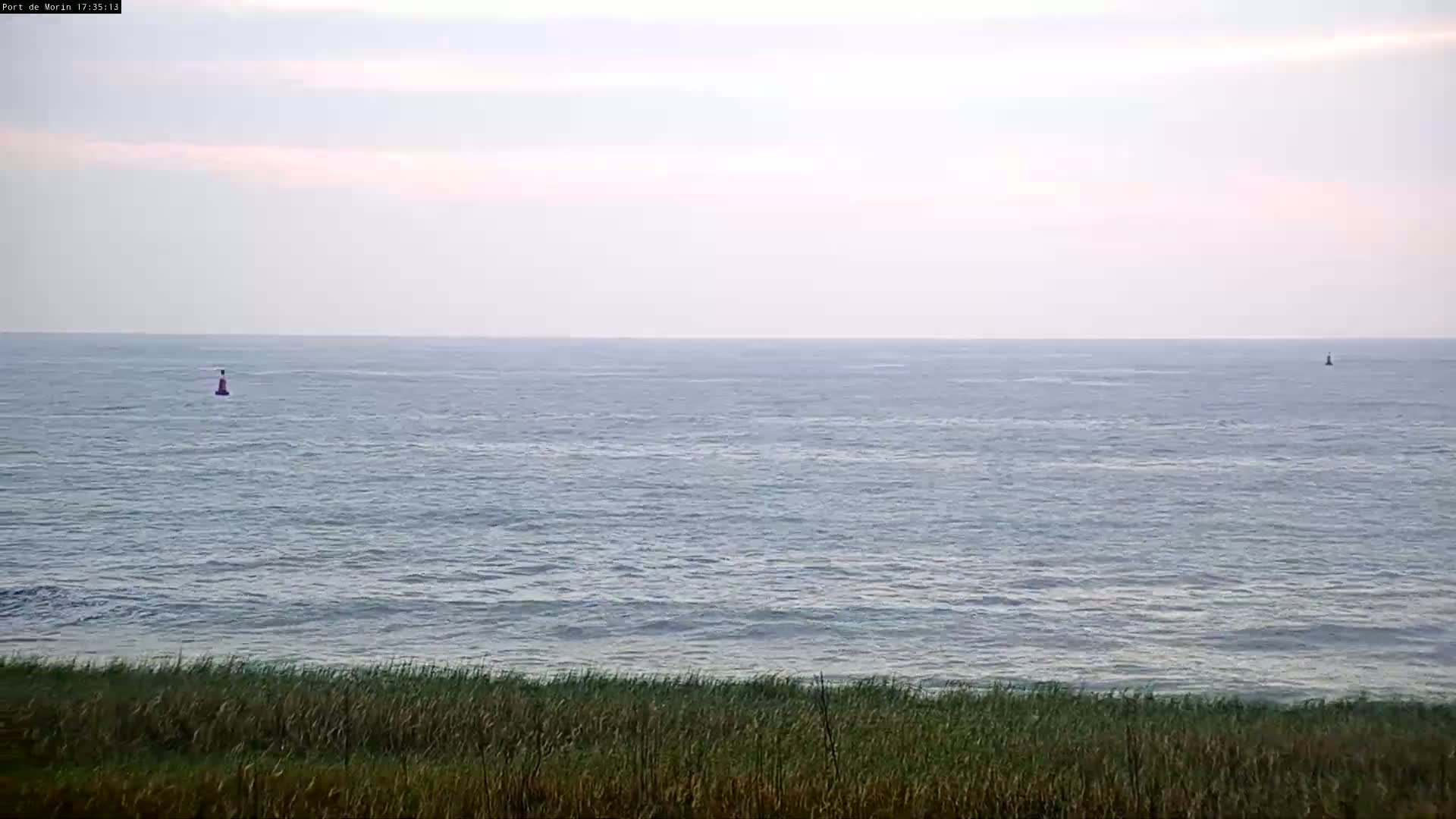 A calm sea with two buoys is visible from a grassy shore under a pale sky.