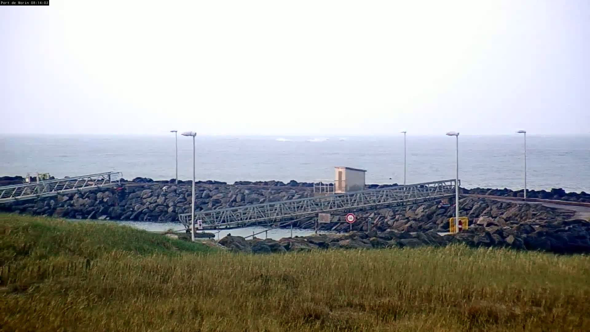 A calm sea is visible beyond a rocky shoreline featuring a small boat launch and several tall lampposts, all observed from a grassy hillside.