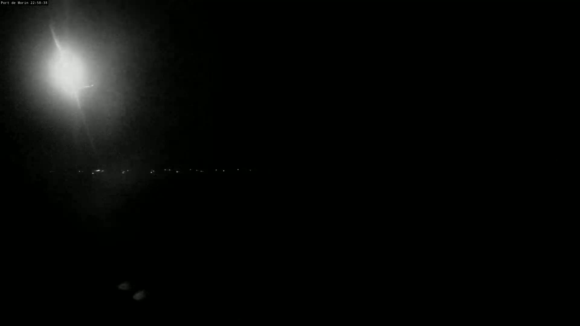 A nighttime, dark scene shows a bright light source in the upper left corner illuminating a distant line of faint lights.