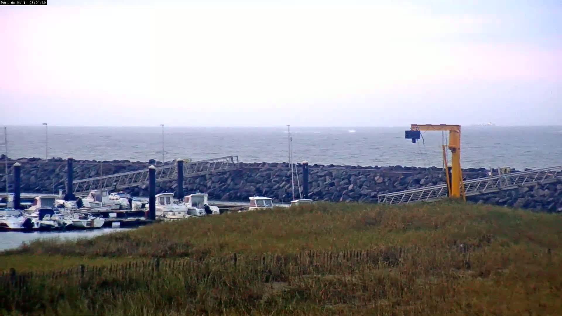 A small harbor with several boats docked behind a low rock wall, a yellow crane, and grassy area, all under a mostly cloudy sky and slightly choppy water.