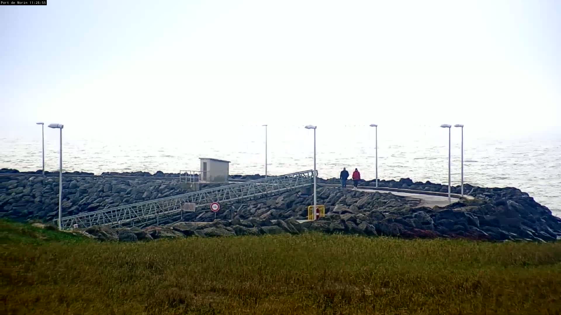 Two people walk along a rocky shoreline with a metal ramp and several lampposts under a hazy, overcast sky and slightly wavy water.