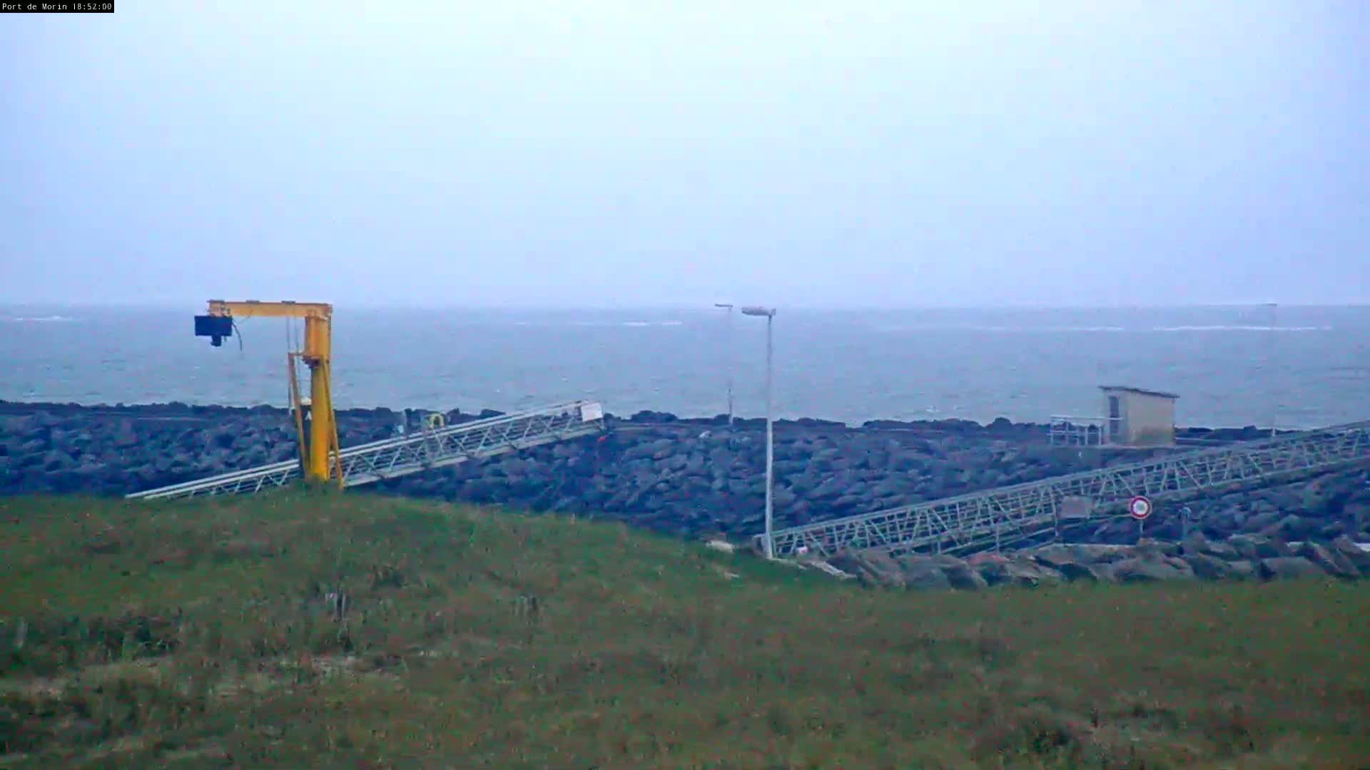 A yellow crane and metal walkways are situated on a grassy hill overlooking a rocky breakwater and a calm, gray sea under an overcast sky.