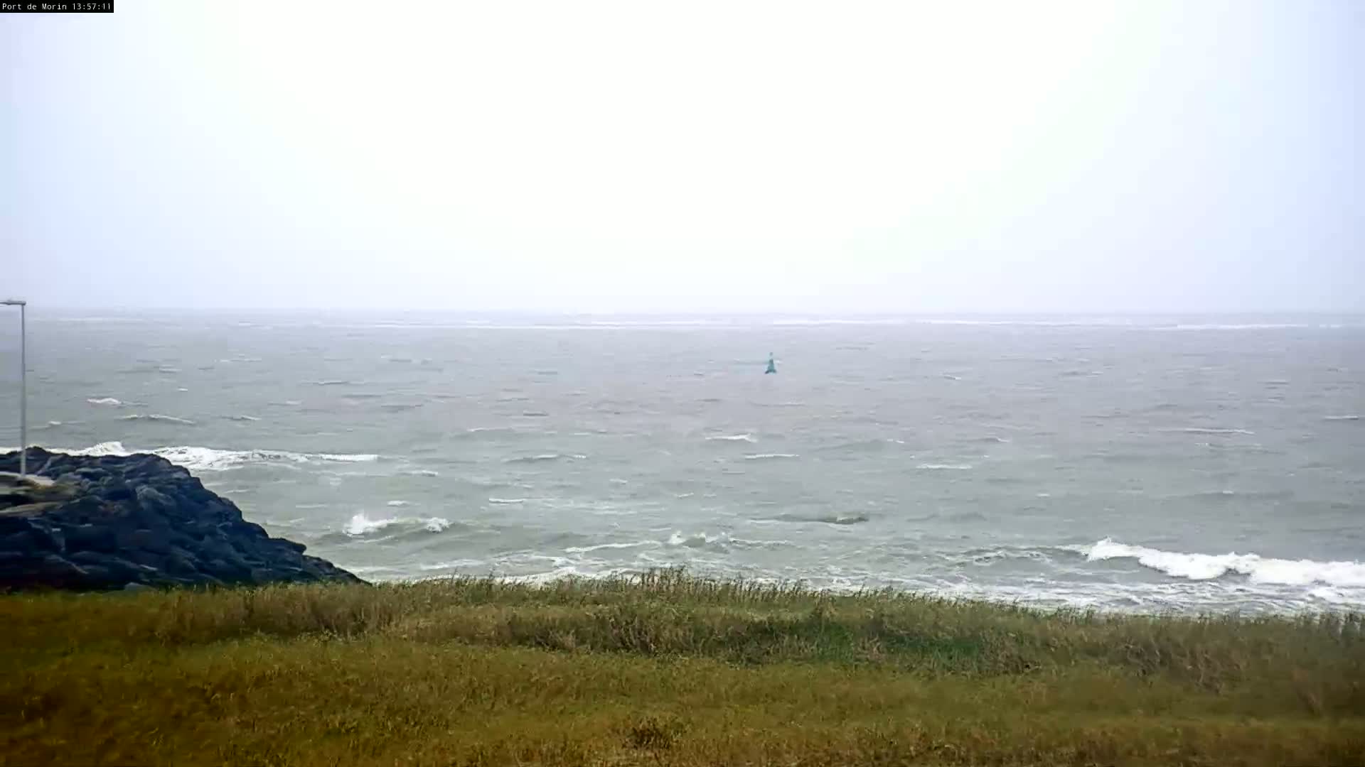 A hazy, overcast day shows a choppy sea with small waves breaking near a rocky shoreline and grassy hill.