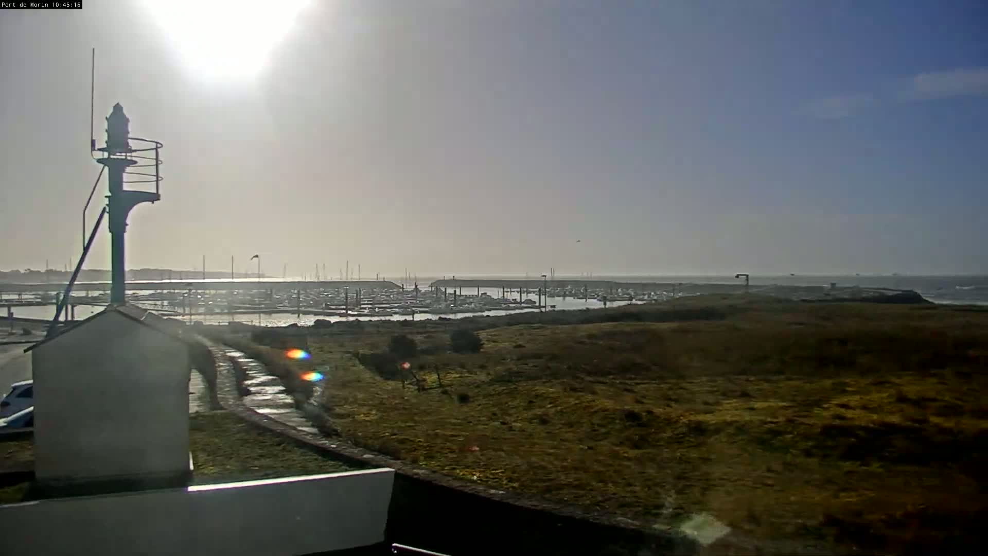 A sunny day reveals a harbor filled with boats, viewed from a grassy hilltop near a small structure and a tall light post.