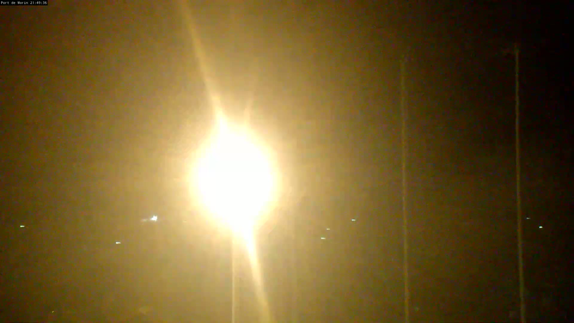 A bright light shines through a hazy, dark night, illuminating indistinct vertical poles and distant faint lights.