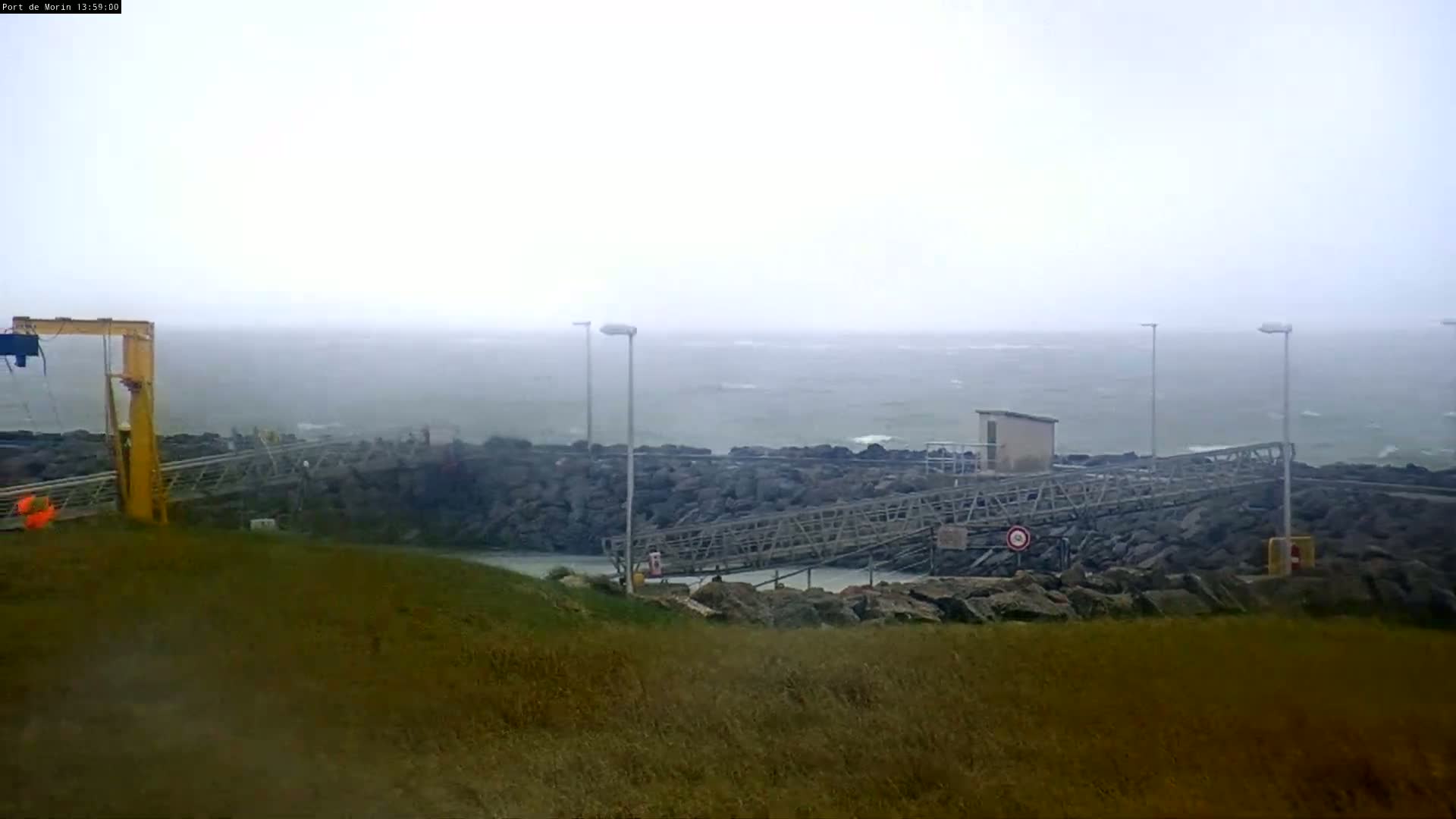 A rocky shoreline with a metal walkway and a yellow crane is visible under overcast skies and windy conditions, with a choppy sea extending into a misty distance.