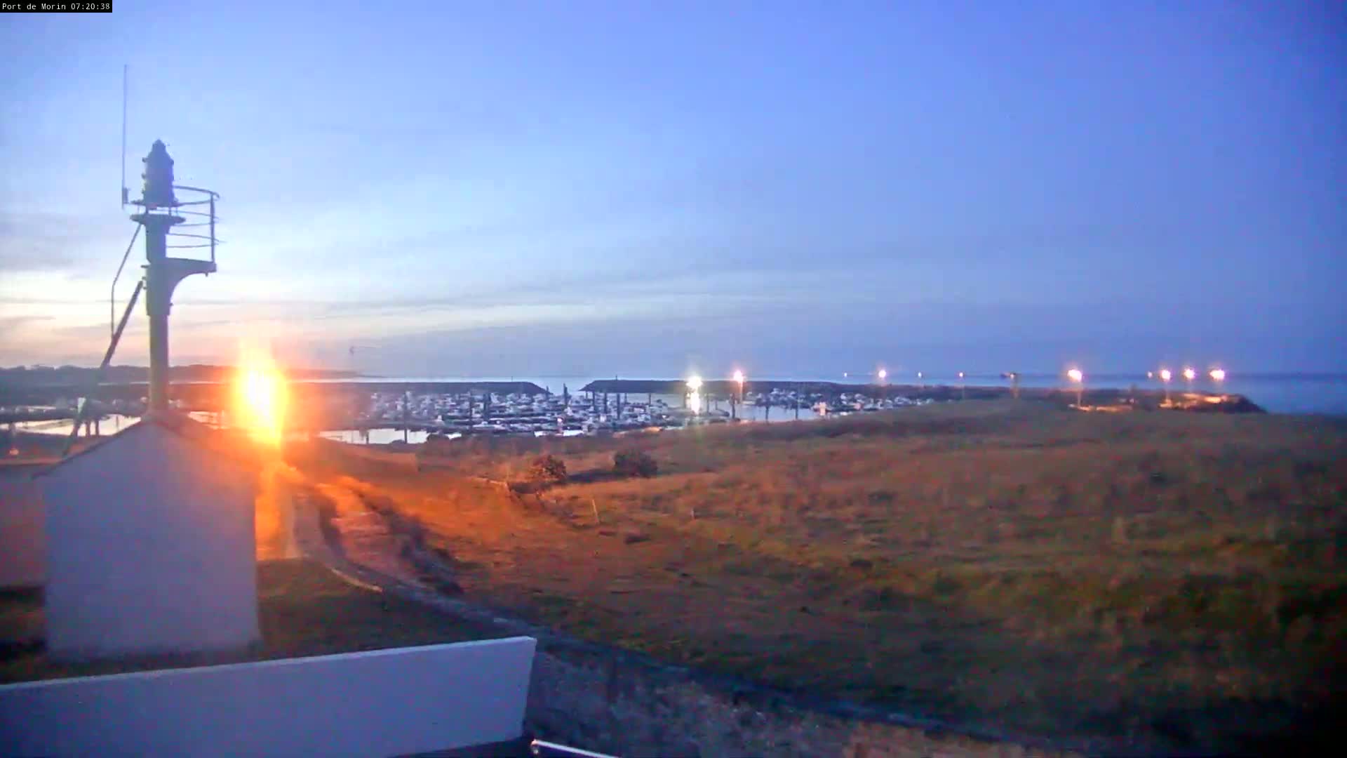 A marina filled with boats is visible at dusk from a vantage point overlooking a grassy area, with a tall light post in the foreground.