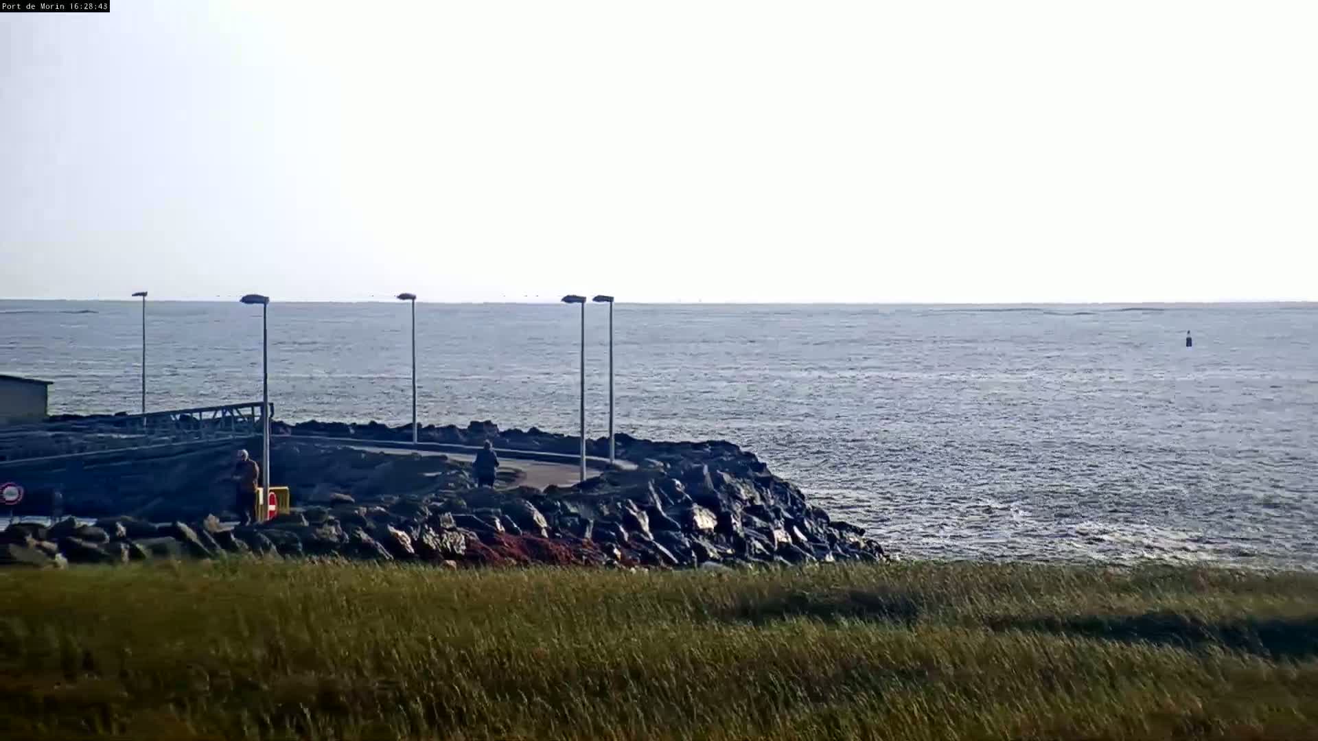 A rocky shoreline with a few people is visible, overlooking a calm sea under a pale sky.