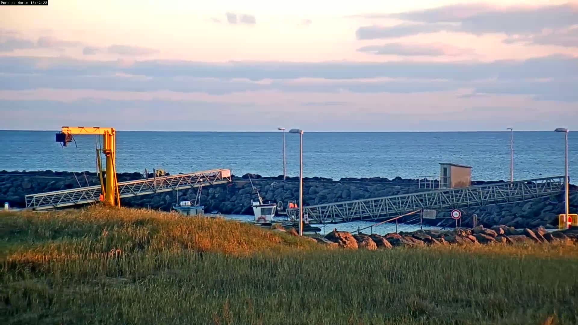 A small harbor with a yellow crane, several boats, and metal walkways sits on a rocky shore next to a calm ocean under a partly cloudy sunset sky.
