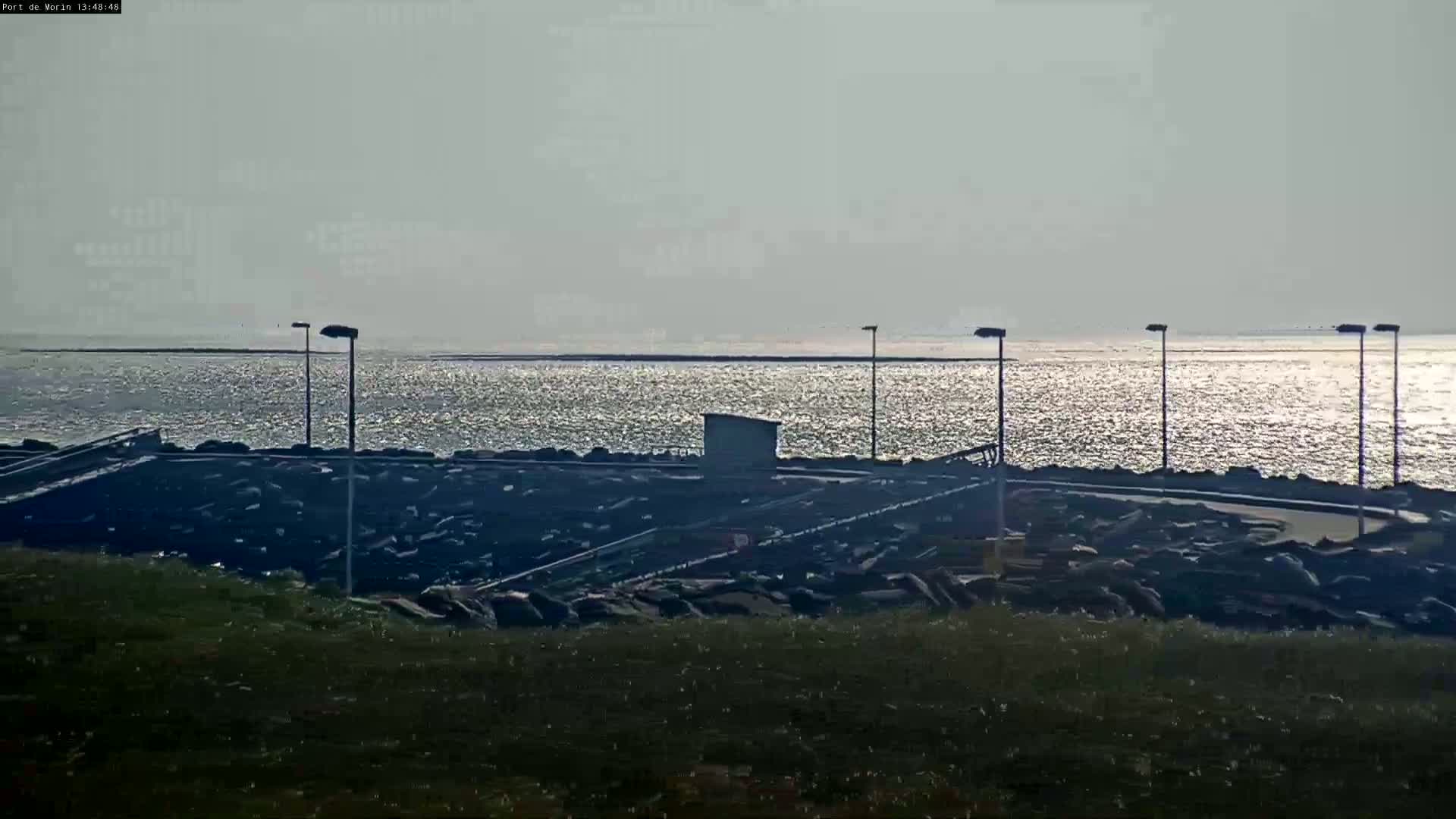A calm, sunlit body of water stretches to the horizon, beyond a rocky shoreline with several lampposts and a small building.