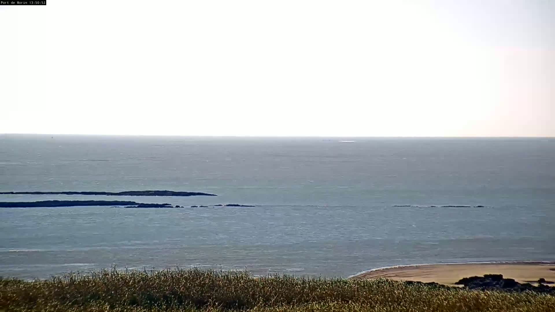 A calm sea with a sandy beach and grassy hill in the foreground under a bright, mostly clear sky.