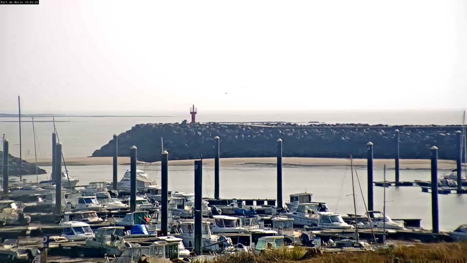 A marina filled with numerous boats is situated near a rocky breakwater, under a hazy sky.