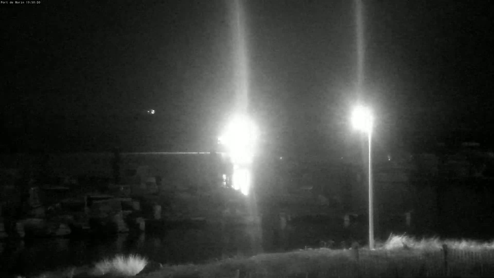 A nighttime, low-light view shows a brightly lit area near a body of water, with several indistinct objects and a tall light post visible in the foreground.