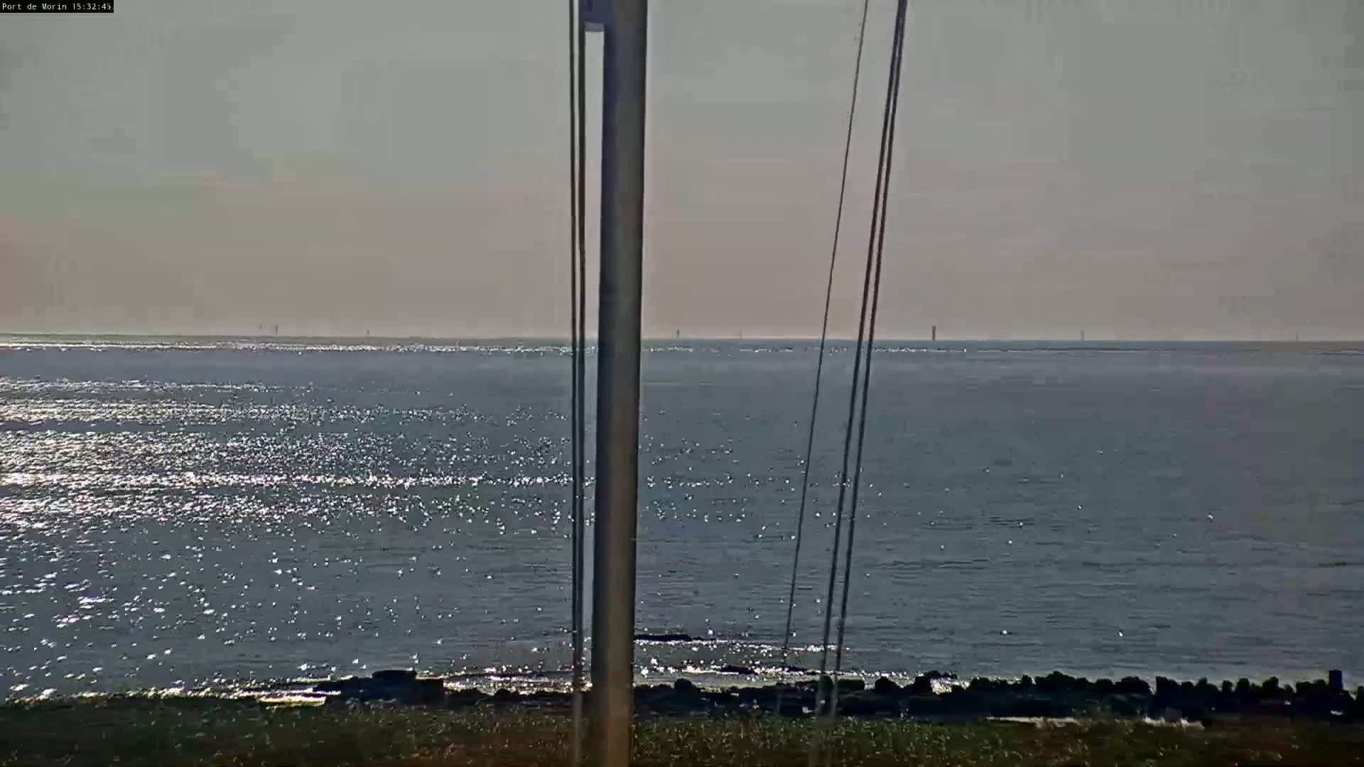 A sparkling body of water stretches to a hazy horizon under a bright, mostly clear sky, viewed from behind a tall post with thin wires.