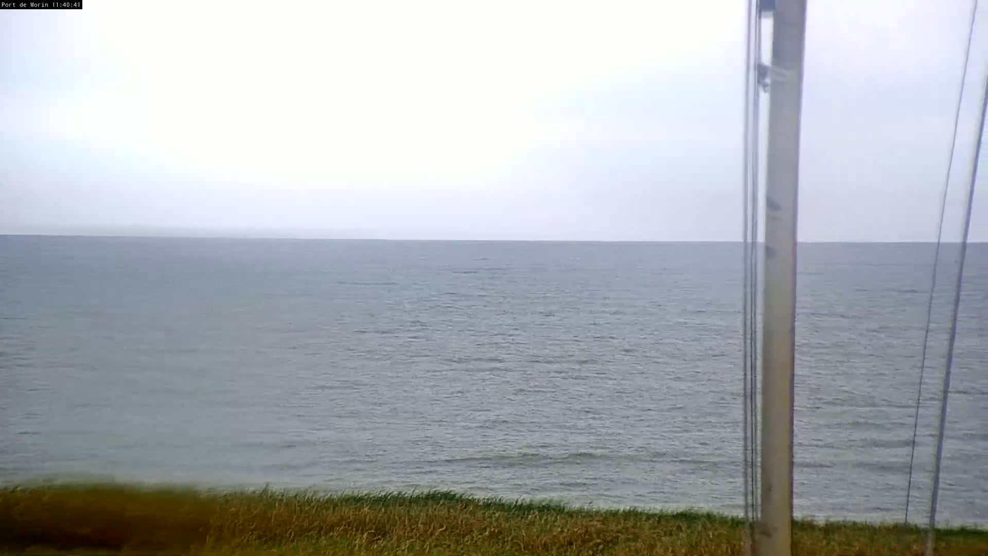 A calm, gray sea stretches to a hazy horizon, viewed from behind tall poles and across a strip of beach grass.