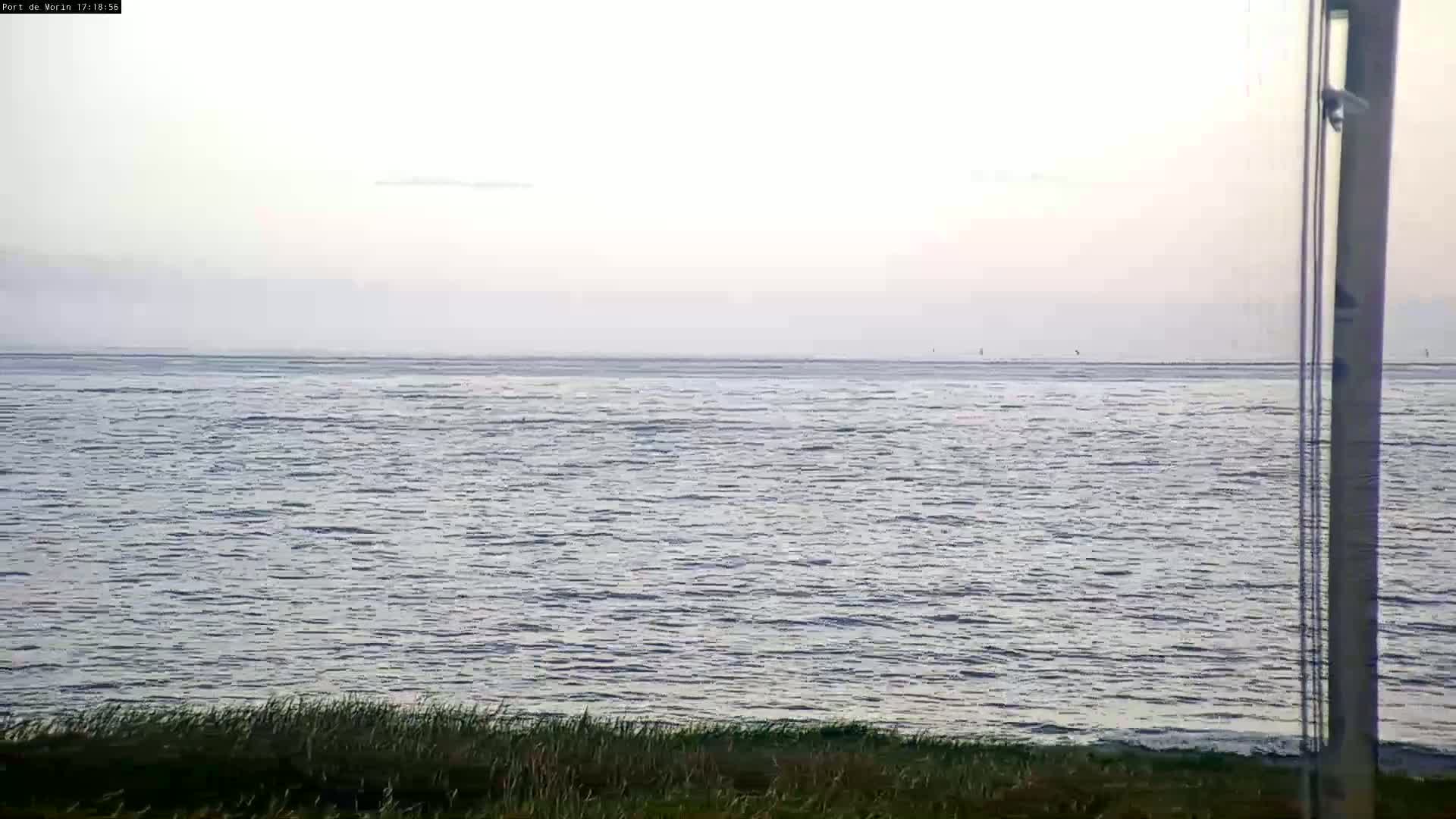 A calm body of water stretches to a hazy horizon under a pale sky, with a strip of grass visible in the foreground.