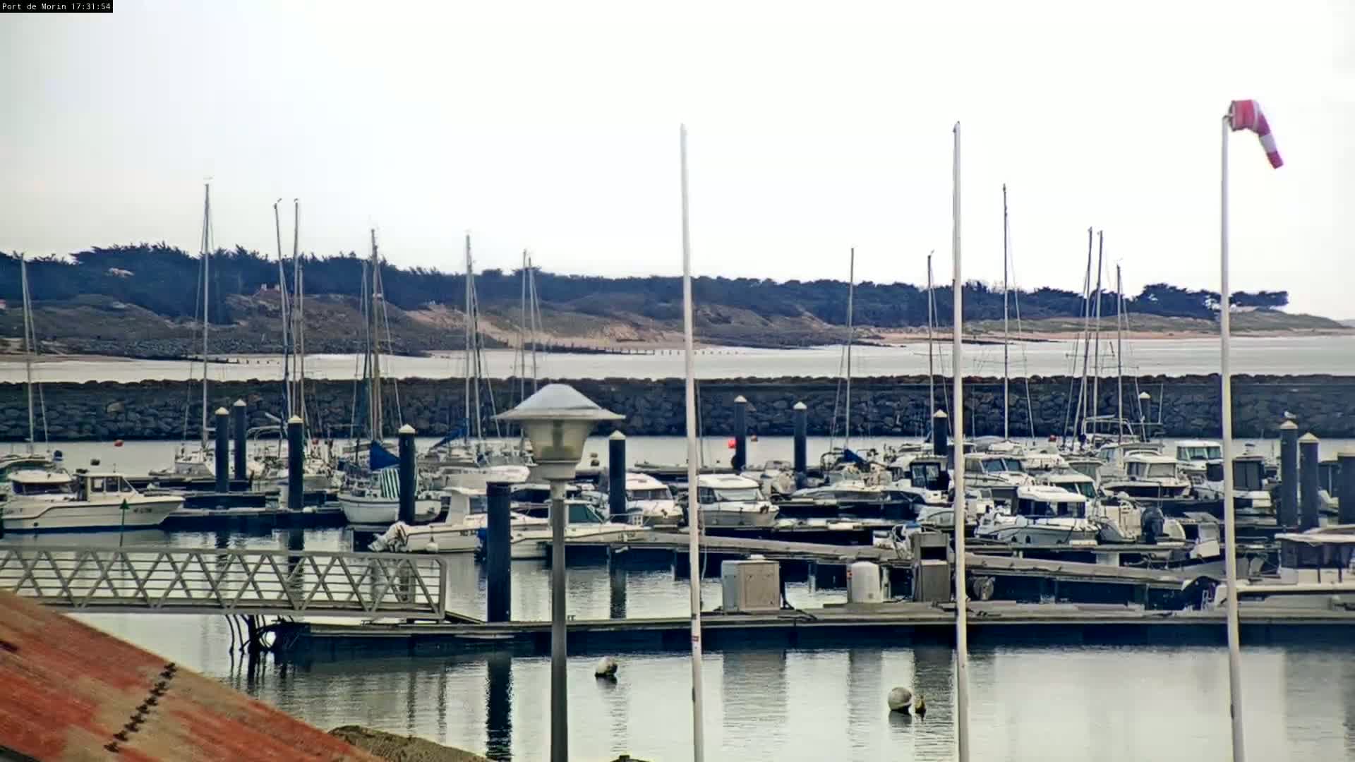 Many boats are docked in a marina on calm water, with a windsock indicating wind and a sandy shoreline visible in the distance under an overcast sky.