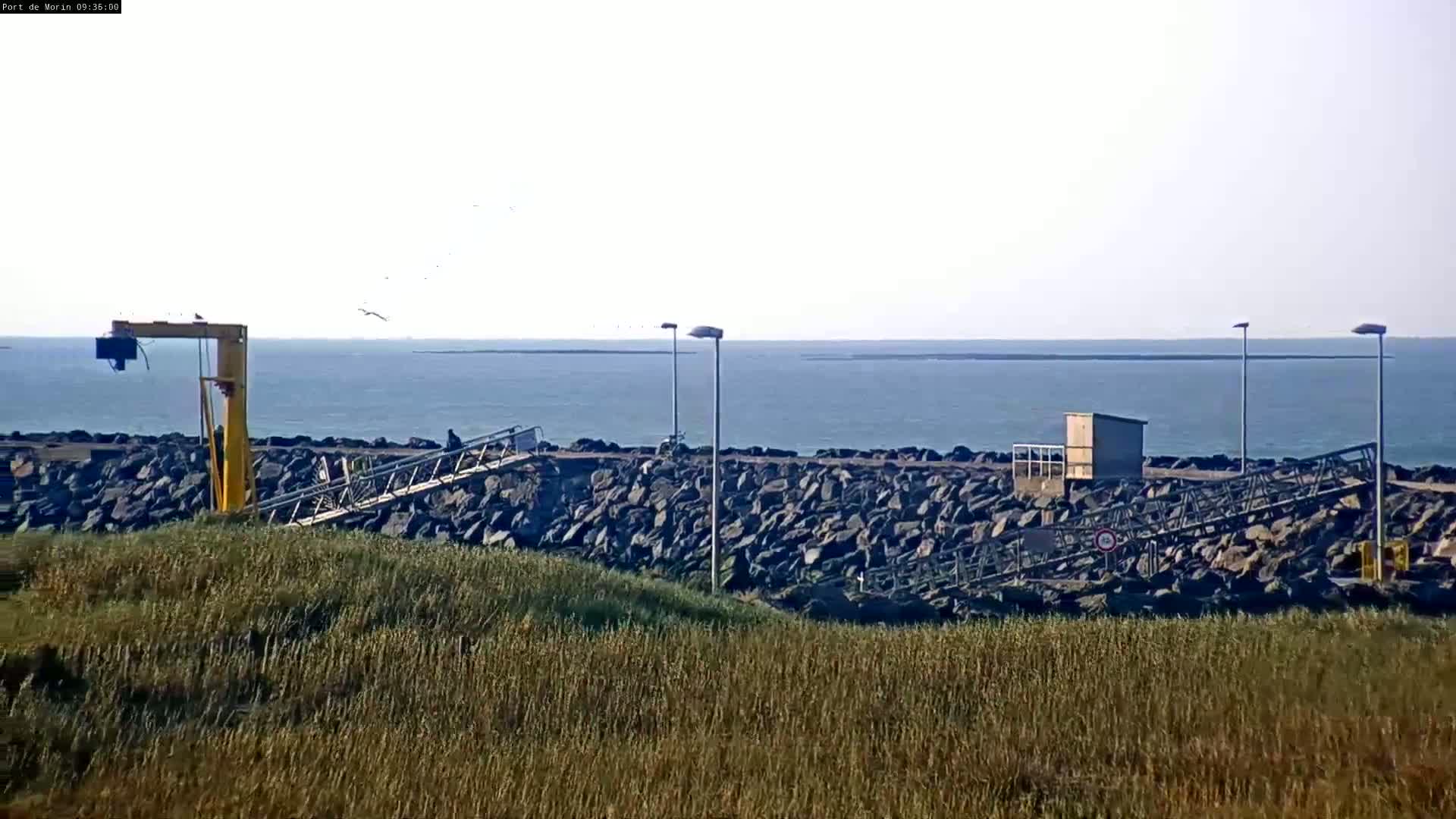 A rocky shoreline with a small pier, a crane, and several lampposts overlooks a calm sea under a clear sky.