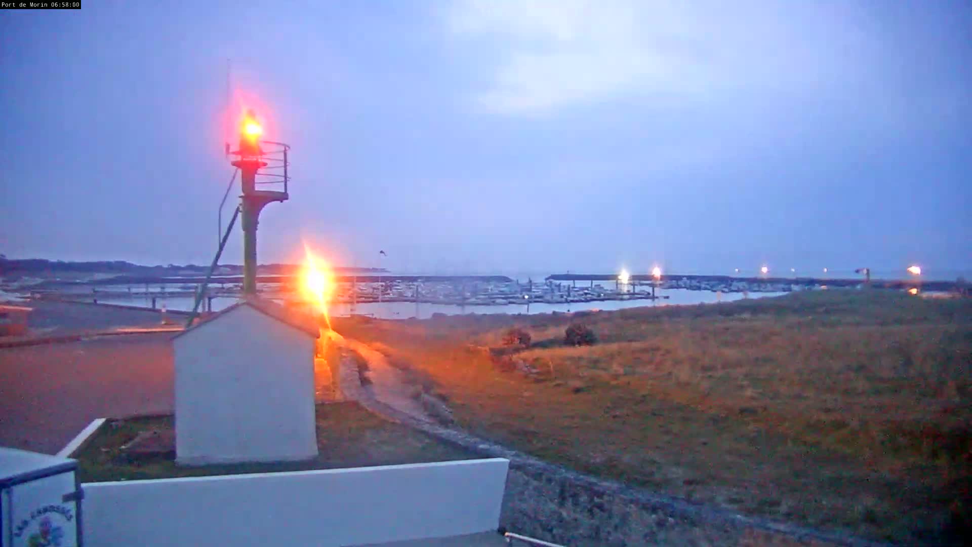 A lit harbor with many boats is visible at dusk from a vantage point near a small, white building and a lit navigation light.