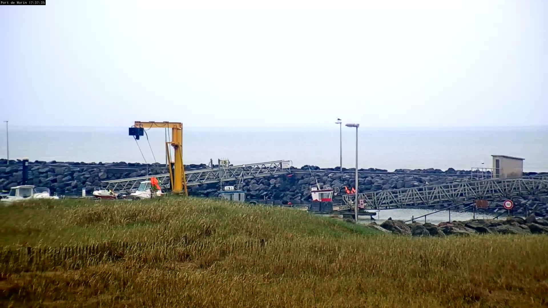A small harbor with a crane and several small boats is visible from a grassy hill overlooking a calm, overcast sea.