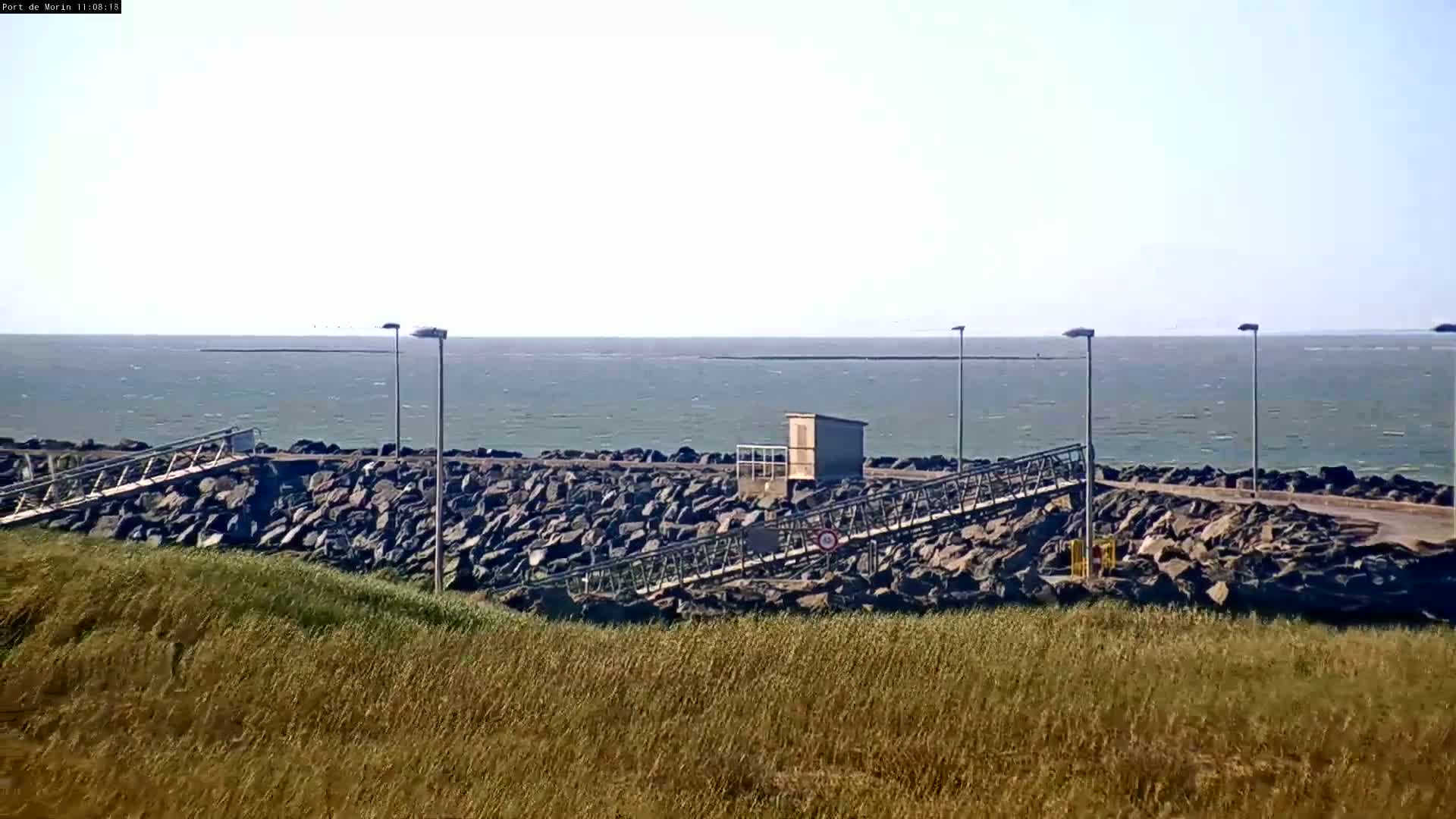 A rocky shoreline with metal walkways and a small building overlooks a calm, gray-green sea under a clear sky.