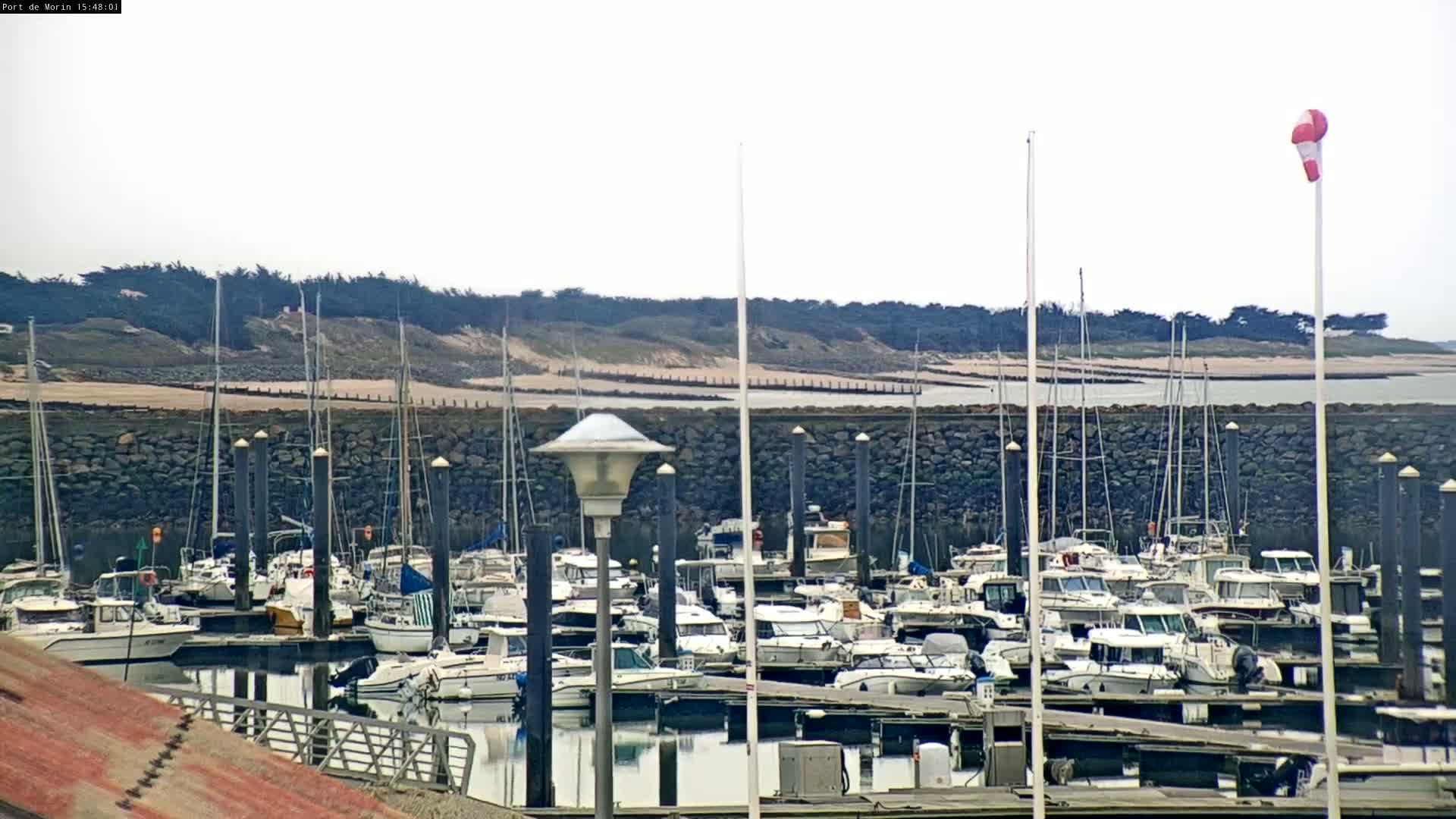 A marina filled with numerous boats is nestled behind a stone wall, with a sandy shore and distant trees visible under an overcast sky.
