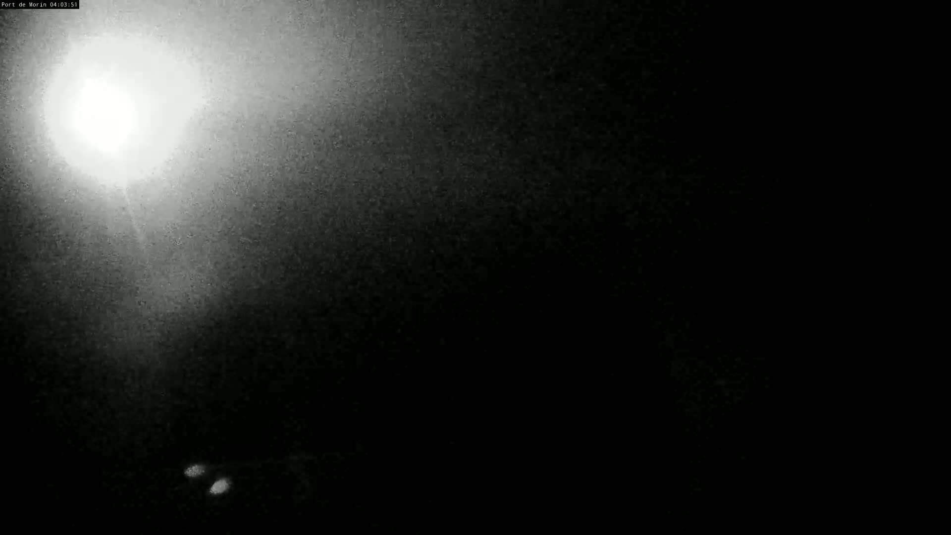 A bright light shines from the upper left corner of a dark, grainy nighttime image, illuminating a small portion of the scene below.